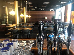 Interior de un restaurante con varias mesas y sillas, una zona de bar con grifos y una pared de madera al fondo.