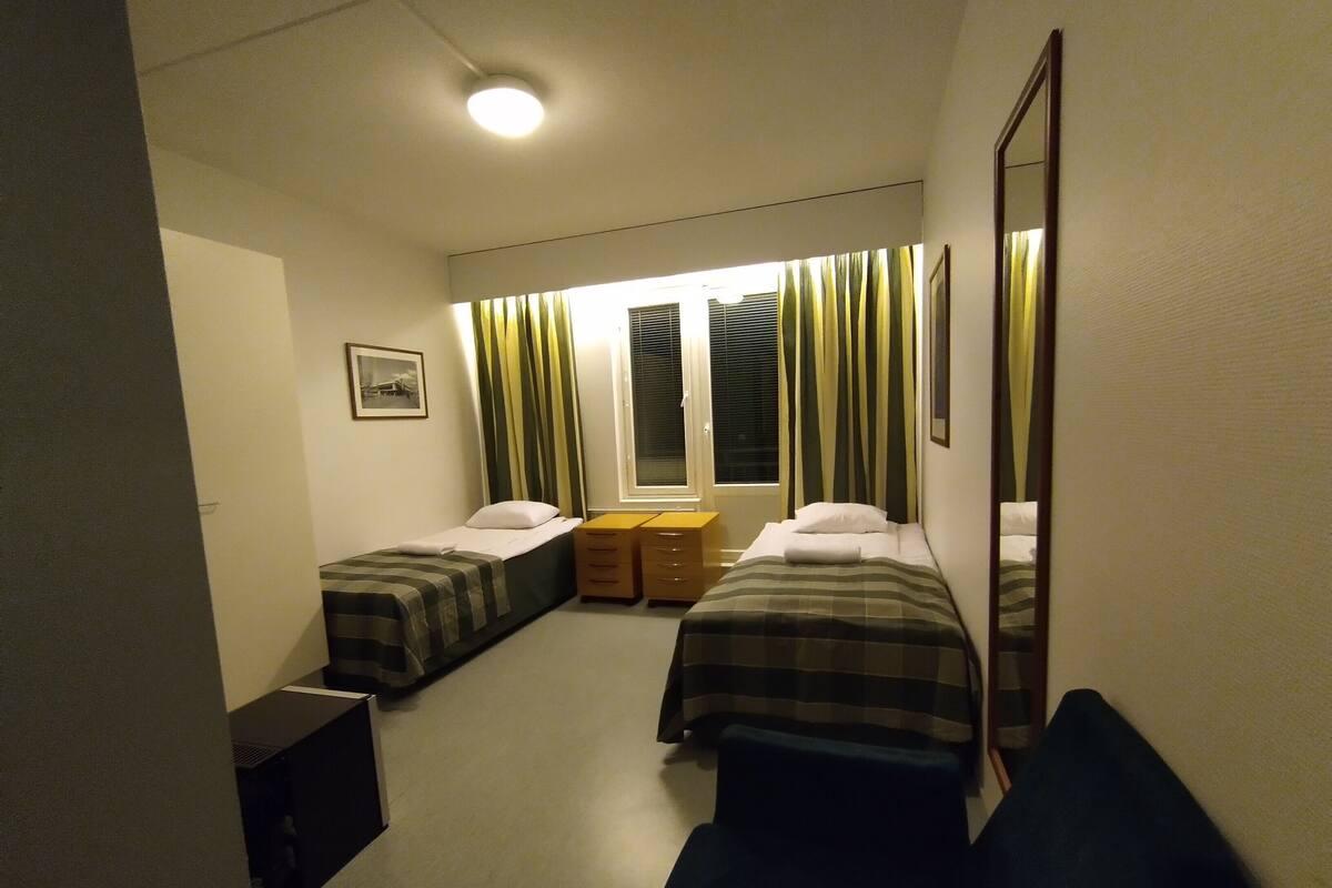 Ett rum med två sängar, ett fönster med gardiner, ett nattduksbord och en spegel.