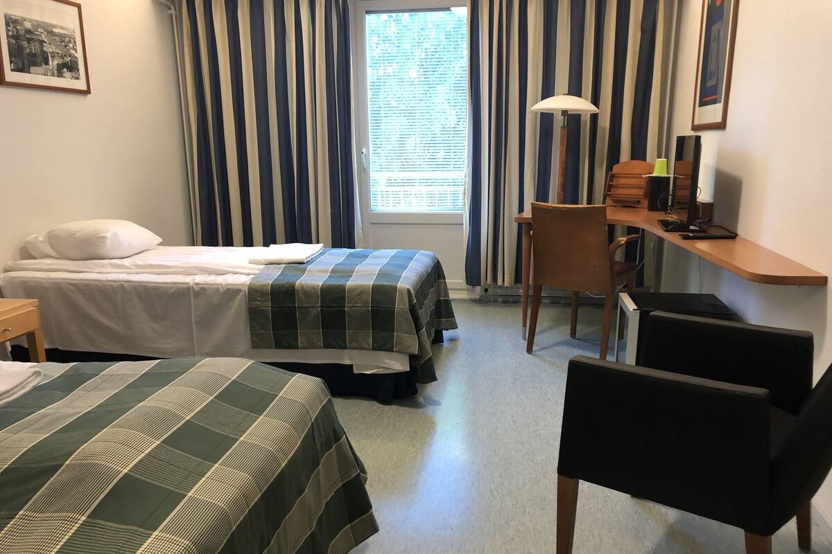Ett hotellrum med två sängar, ett skrivbord med en stol, en lampa och ett fönster med gardiner.