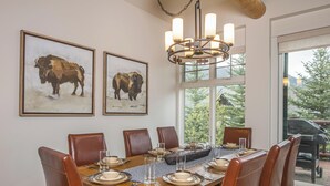 Ein Esszimmer mit einem Holztisch, gedeckt für sechs Personen, umgeben von Ledersesseln. An der Wand befinden sich zwei gerahmte Bison-Gemälde, und eine Chandelier hängt darüber.