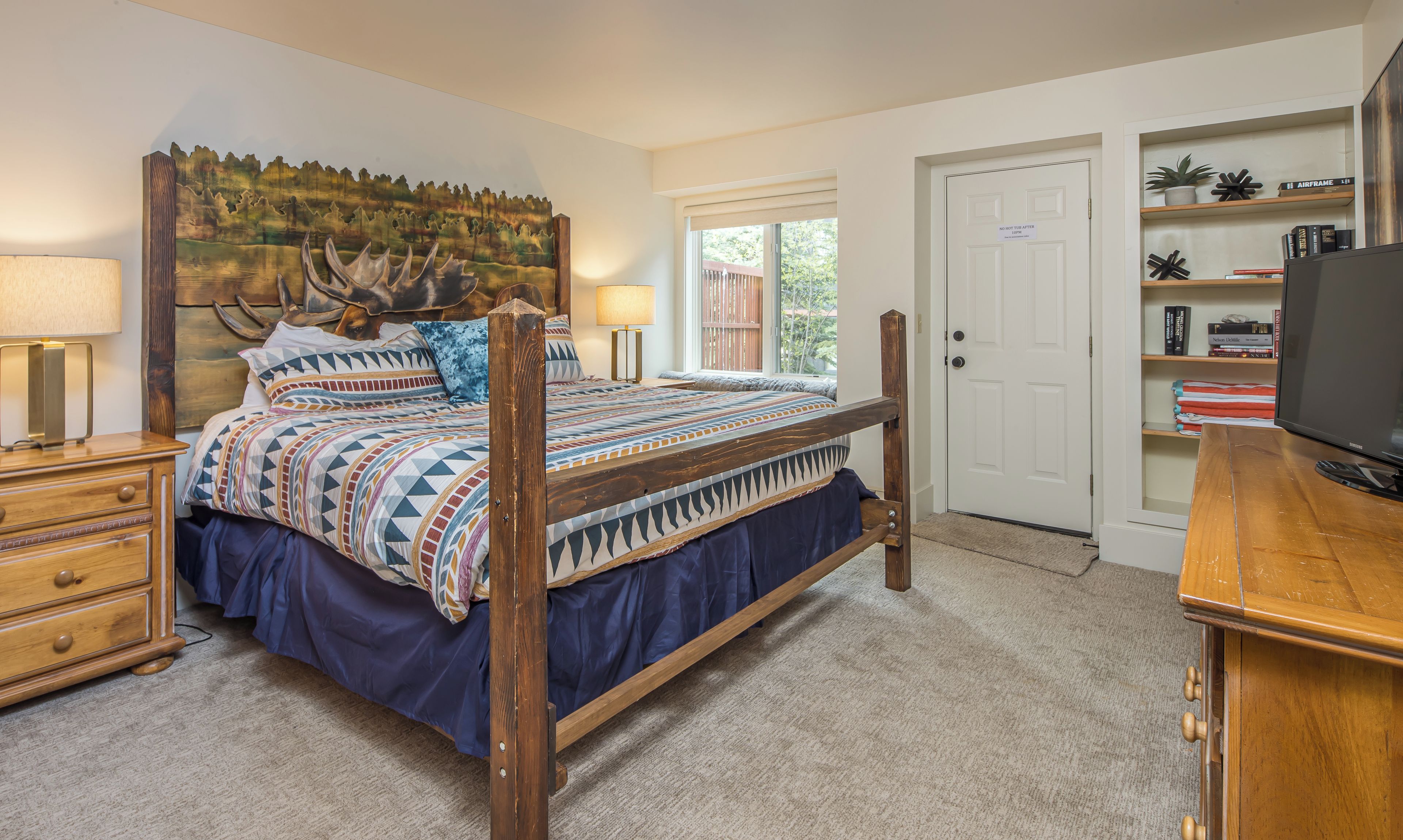 A bedroom with a wooden bed, a headboard with a deer head, a window, a door, and a bookshelf with books and decorative items.