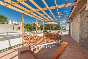 A covered outdoor patio with a hammock, wooden table, chairs, and a white umbrella. - Relax in this modern flat in Baska on Krk (Baska)