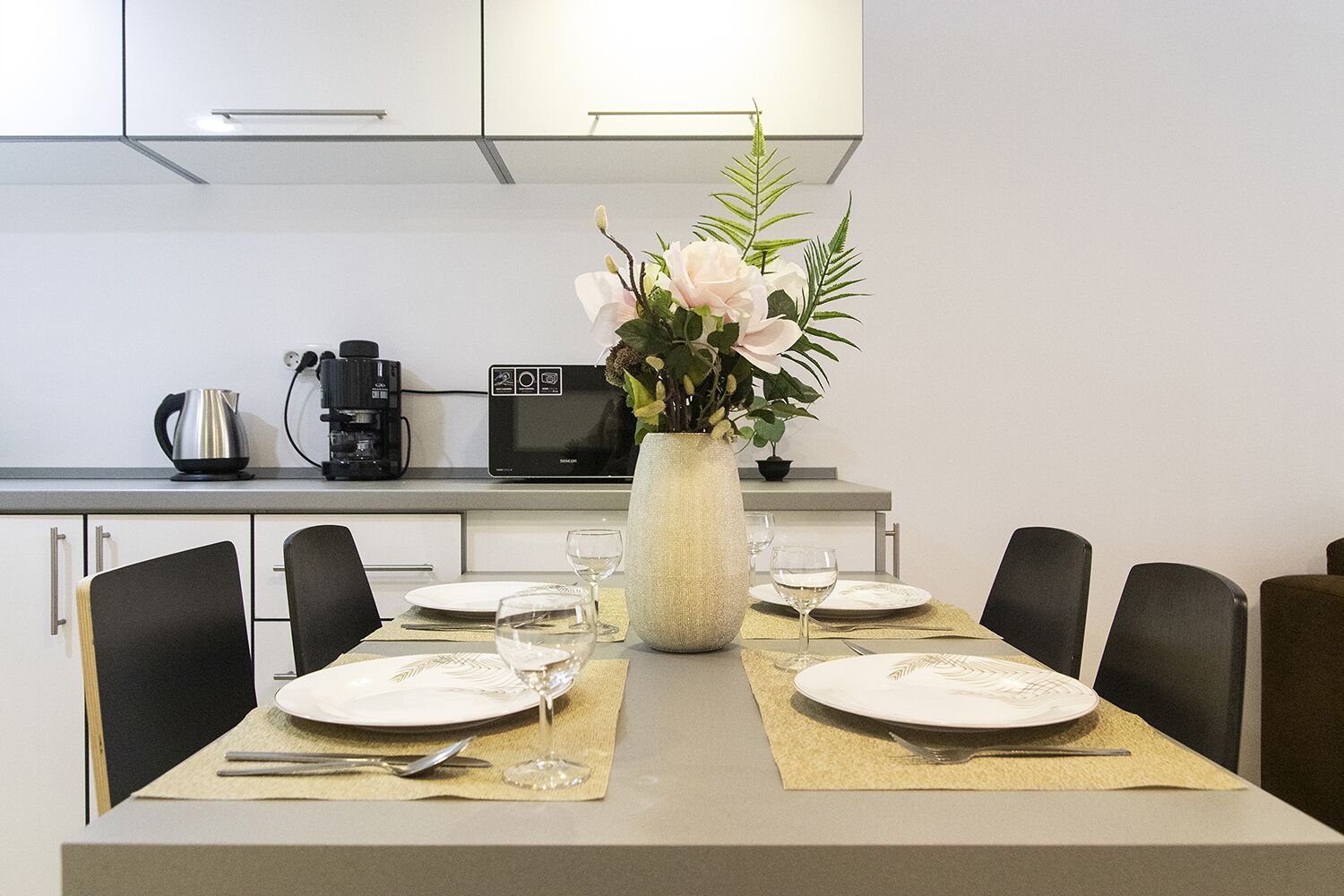 A dining table set with plates, cutlery, and wine glasses, centered with a vase of flowers.