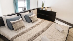 Cama bem arrumada com colcha estampada e travesseiros, mesa de cabeceira com uma planta e um objeto emoldurado, e vista do exterior através de uma janela com cortinas.