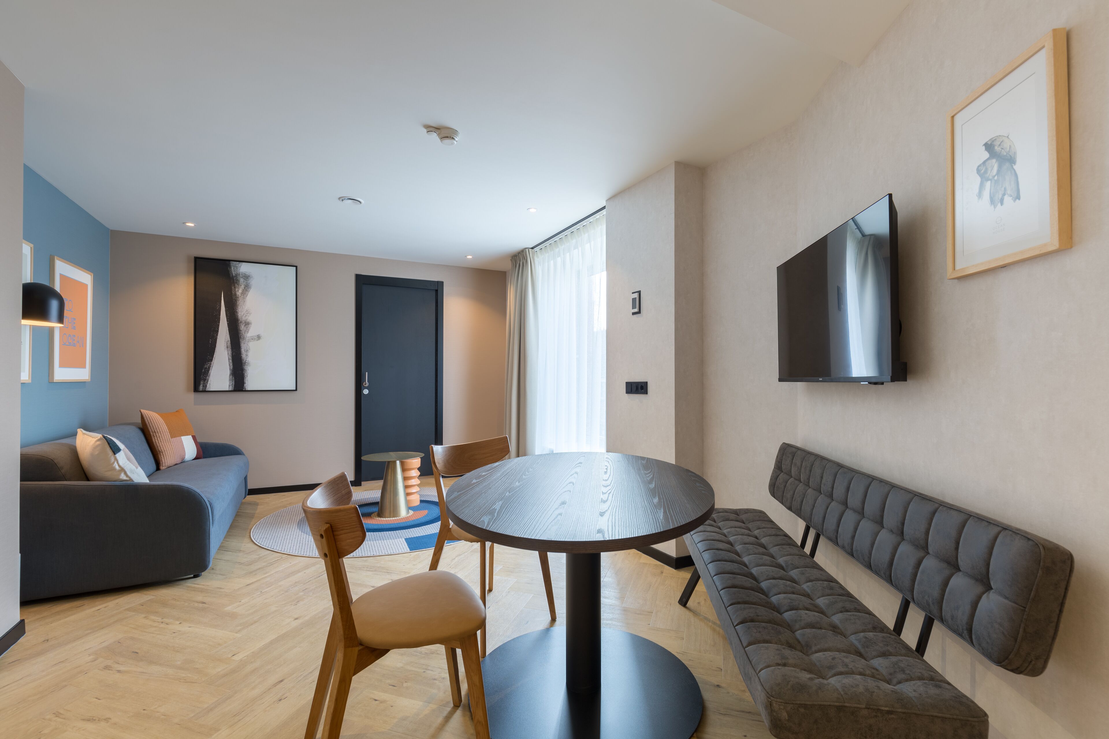 Un salon moderne avec une table ronde, un canapé, un banc et un téléviseur fixé au mur.