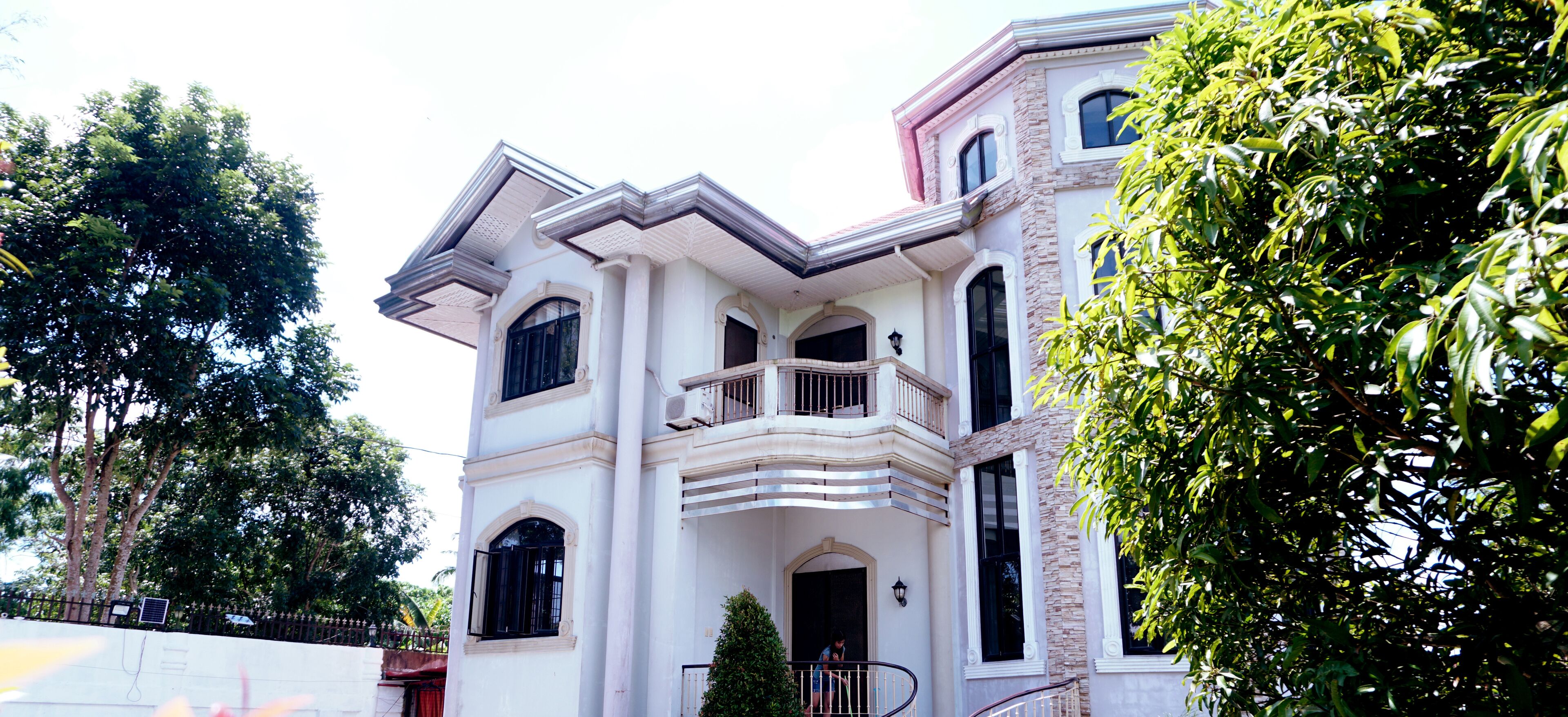 A large, multi-story house with a balcony, surrounded by trees.