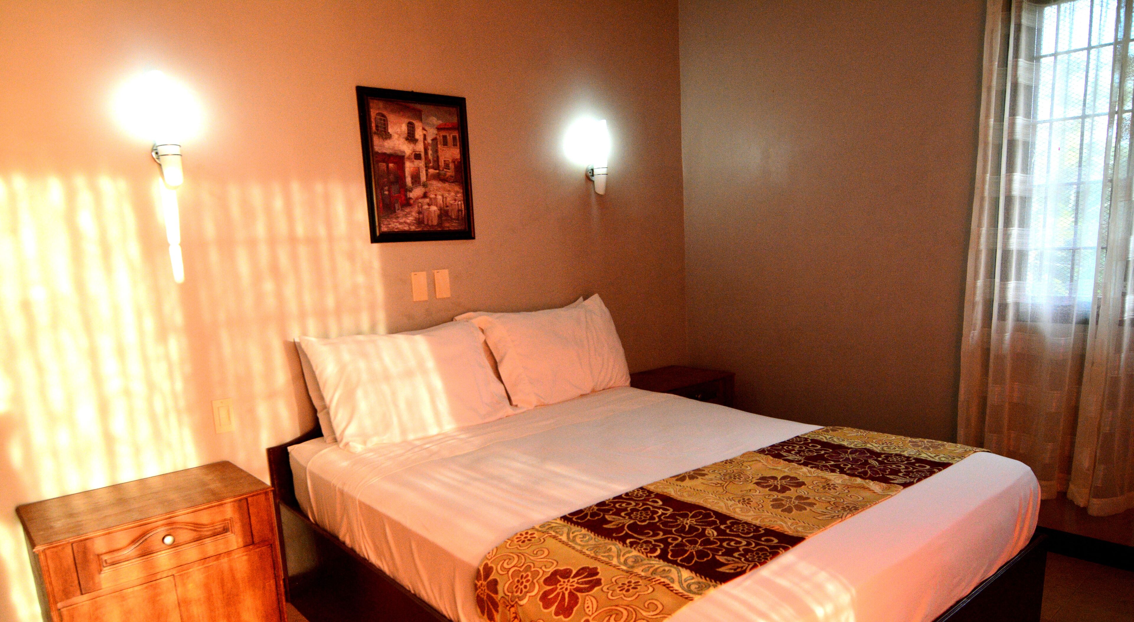 Habitación de hotel con una cama, una mesita de noche, un cuadro colgado en la pared y dos apliques.