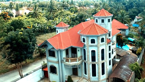 A large, multi-story building with a red-tiled roof, surrounded by lush greenery and palm trees.