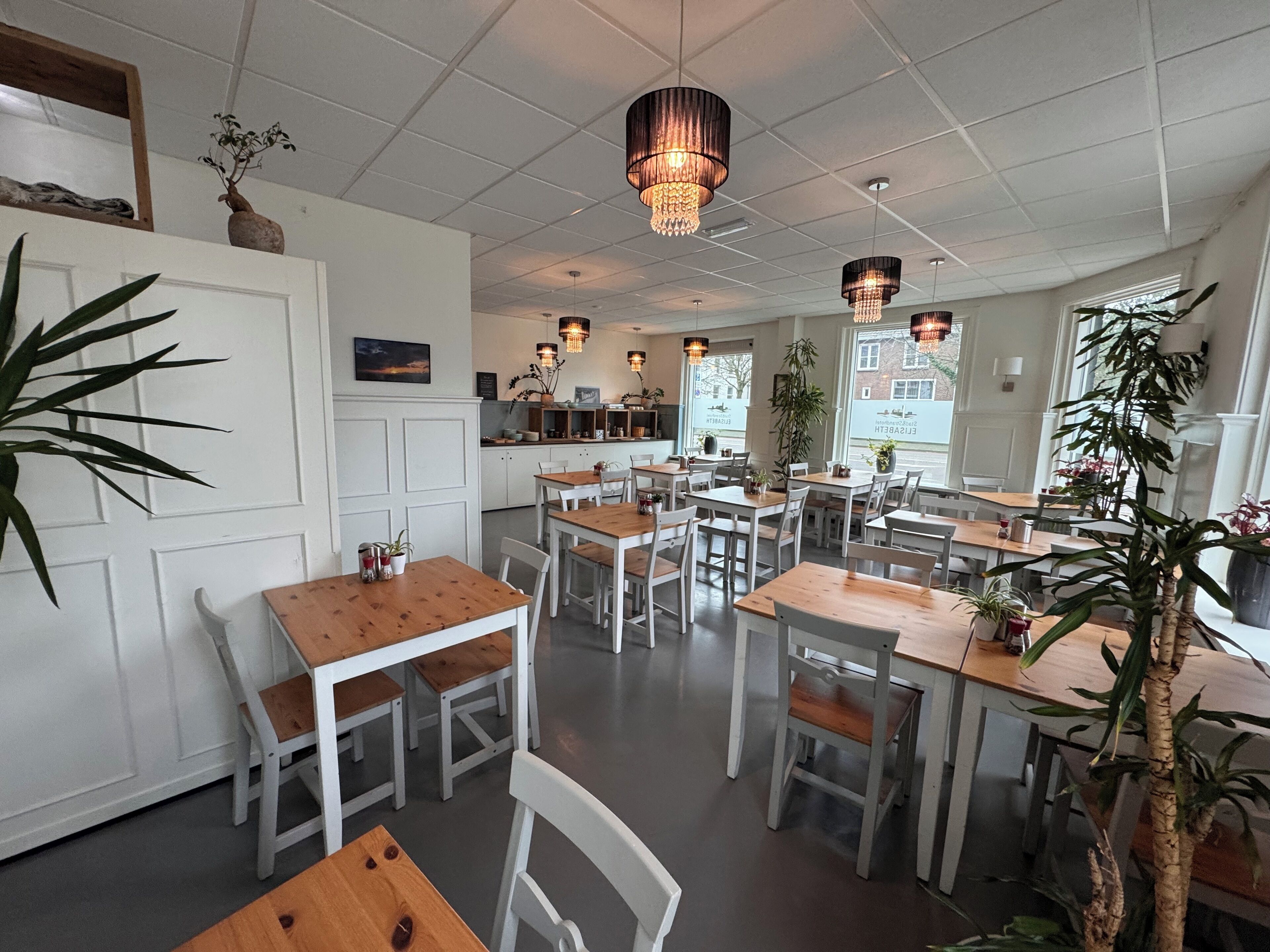 Un restaurant spacieux aux murs blancs, avec des tables et des chaises en bois. On y trouve des lustres suspendus et des plantes en pot.