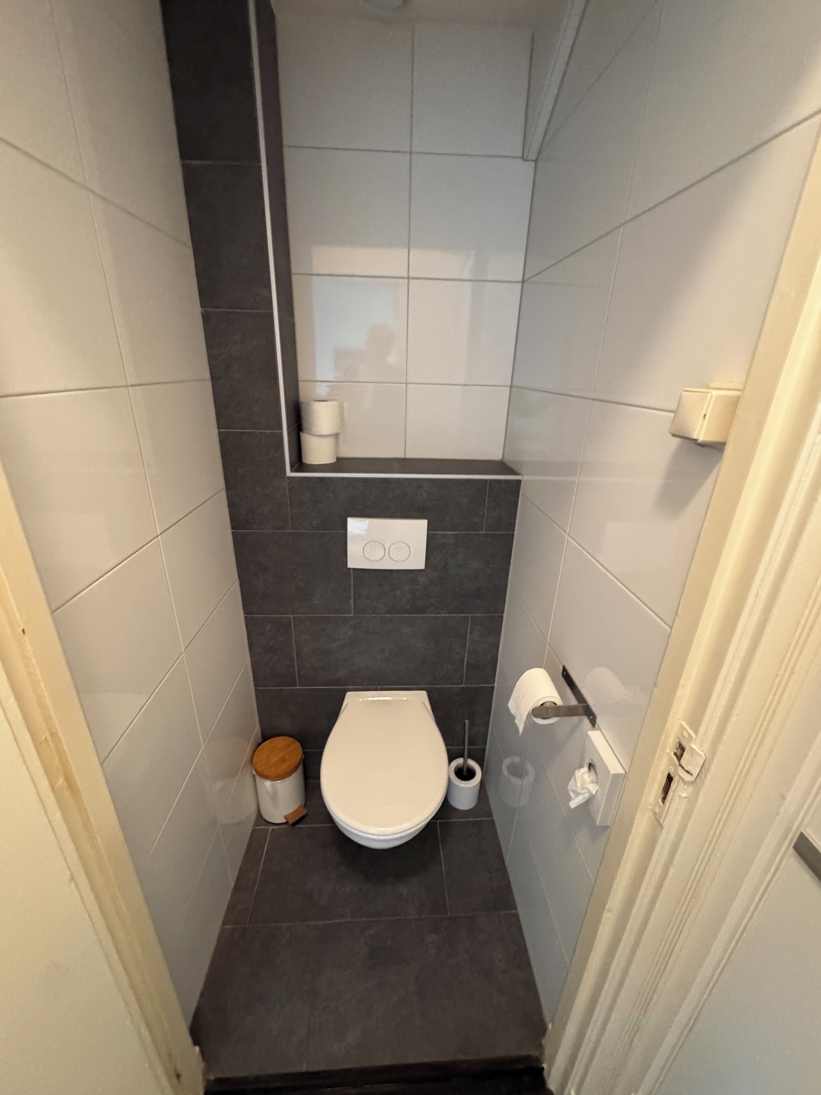 Un baño pequeño con inodoro, un cubo de basura, un porta papel higiénico y un espejo.