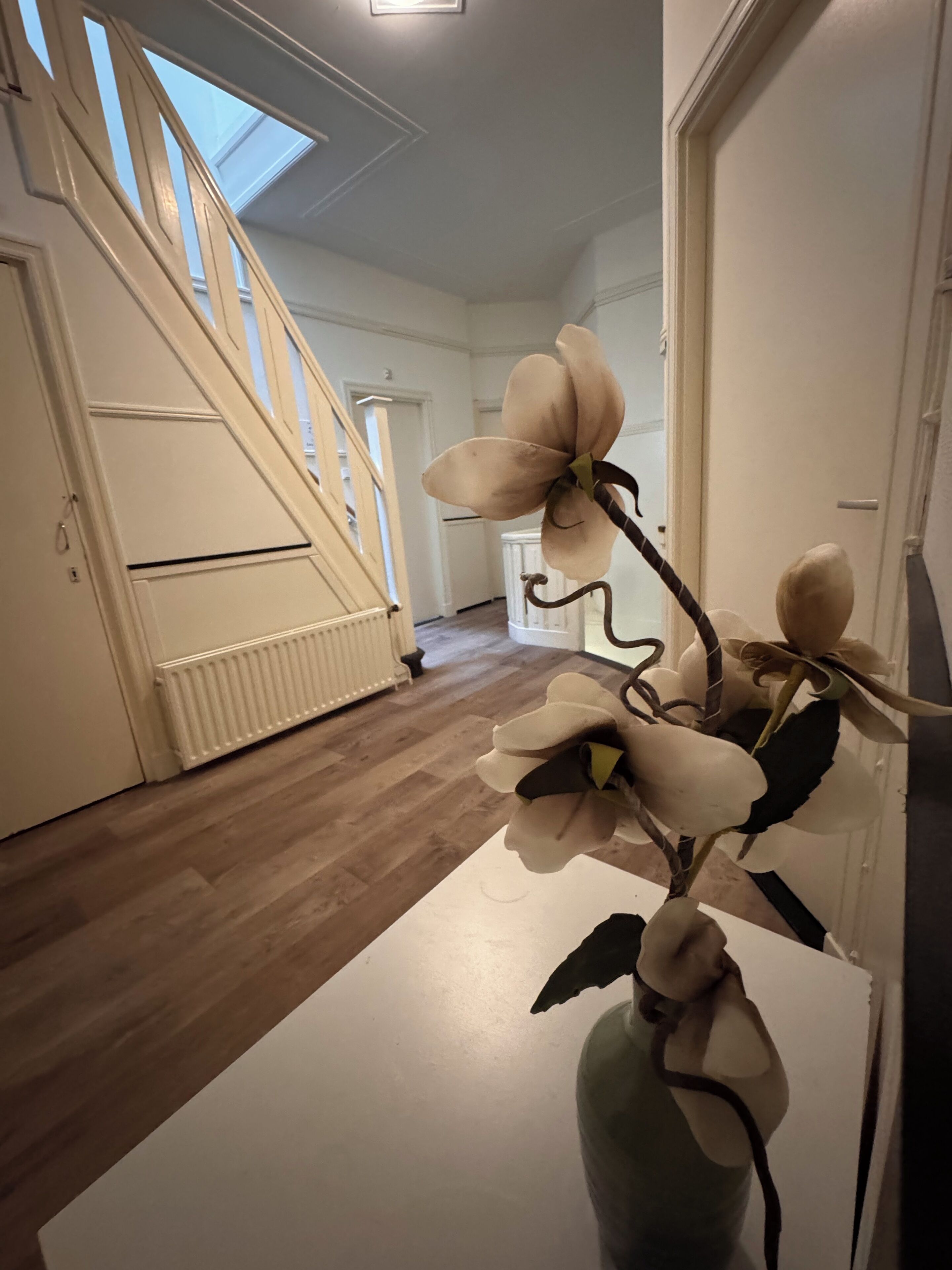 A hallway with a wooden staircase, a white radiator, and a large white flower in a vase.