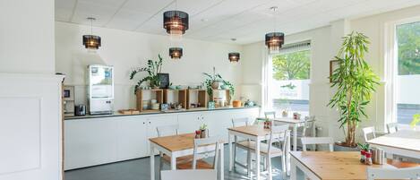 Una cocina moderna con armarios blancos, encimeras de madera e iluminación colgante. Hay varias mesas y sillas, y un ventanal aporta luz natural.