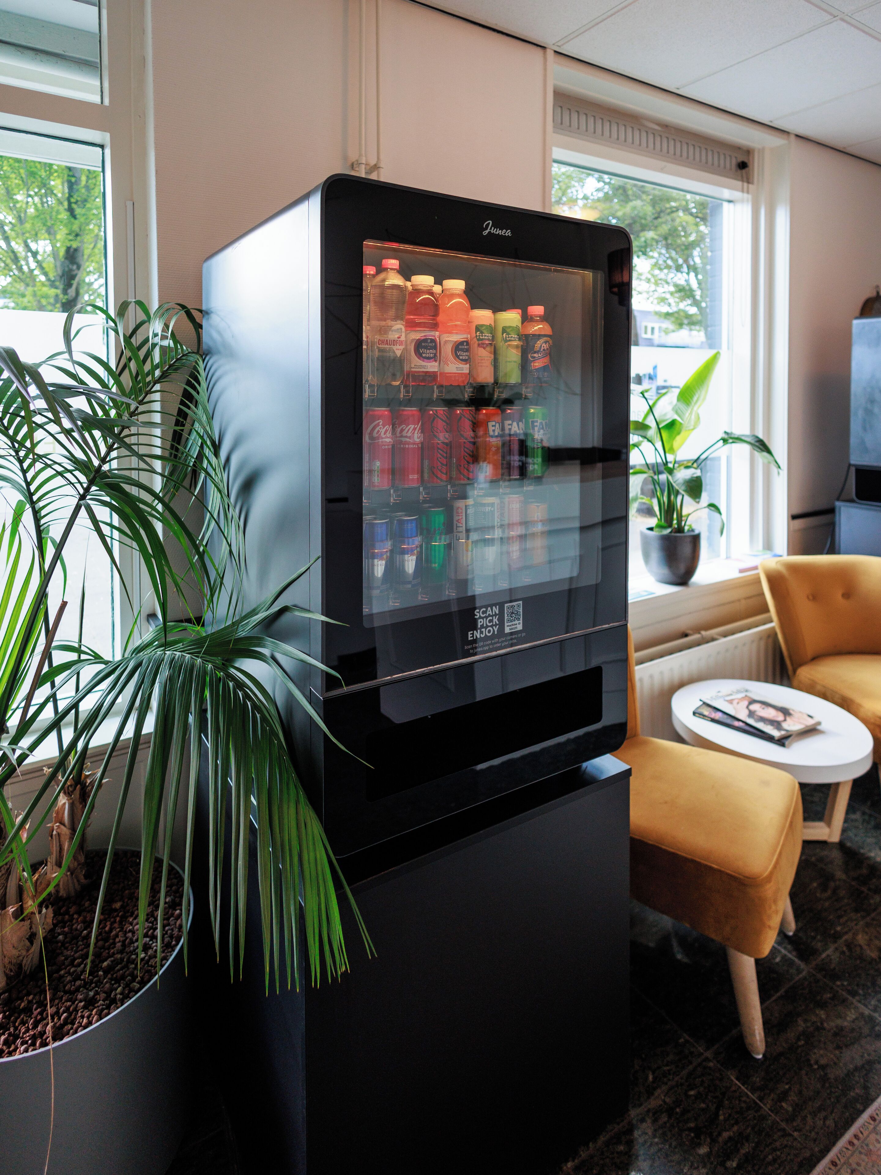 A vending machine with a variety of drinks, a potted plant, a small table with a magazine, and a yellow chair in a room with windows.