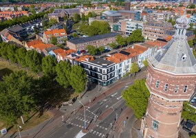 Vista aérea de un paisaje urbano con una torre de ladrillo rojo destacada, una mezcla de edificios tradicionales y modernos, y una calle central con pasos de cebra.