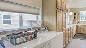 Une buanderie équipée d’un lave-linge et d’un sèche-linge à chargement frontal, de meubles en bois, d’une fenêtre avec stores et d’un coin cuisine avec une cuisinière et des placards.