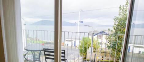 Un balcón con una mesa y sillas, con vista a una calle con edificios y una montaña al fondo.