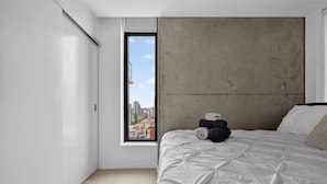 A minimalist bedroom with a large window, a bed with a white duvet, and a concrete accent wall. - King Bed | DT Views | Mins to Stampede | Gym | AC (Calgary)