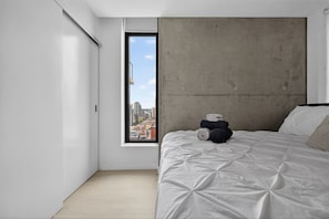 Quarto minimalista com uma janela grande, uma cama com edredom branco e uma parede de destaque em concreto.
