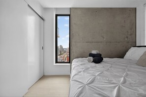 Une chambre à coucher au style minimaliste, avec une grande fenêtre, un mur d’accent en béton et un lit parfaitement fait.