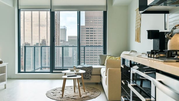 A modern apartment with a large window, a kitchen with a stove, a sofa, and a small round table. - King Bed | DT Views | Mins to Stampede | Gym | AC (Calgary)