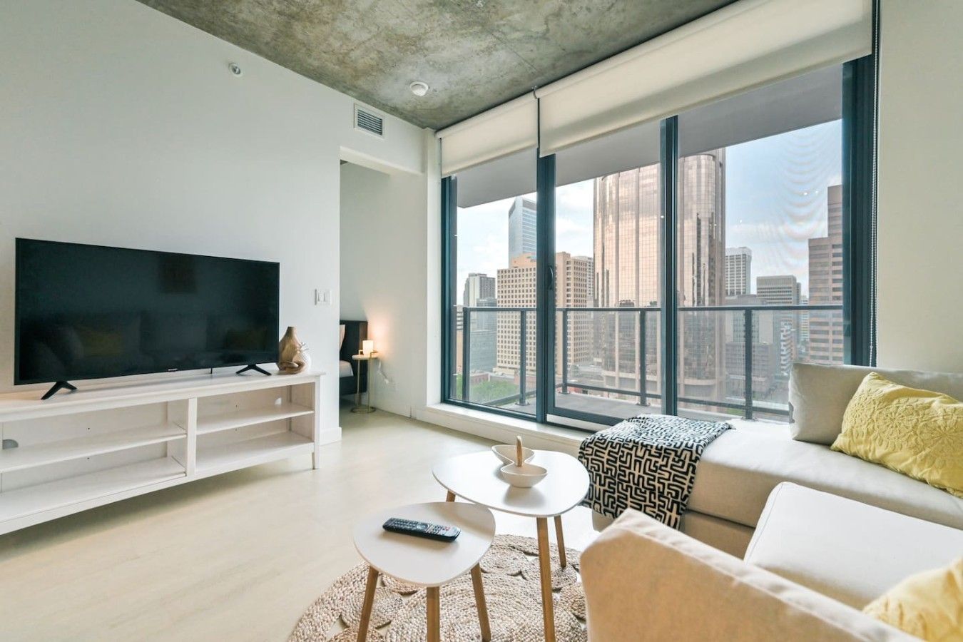 A modern living room with a large flat-screen TV, a sofa, a coffee table, and a view of a cityscape through large windows.