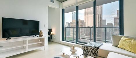 A modern living room with a large flat-screen TV, a sofa, a coffee table, and a view of a cityscape through large windows.