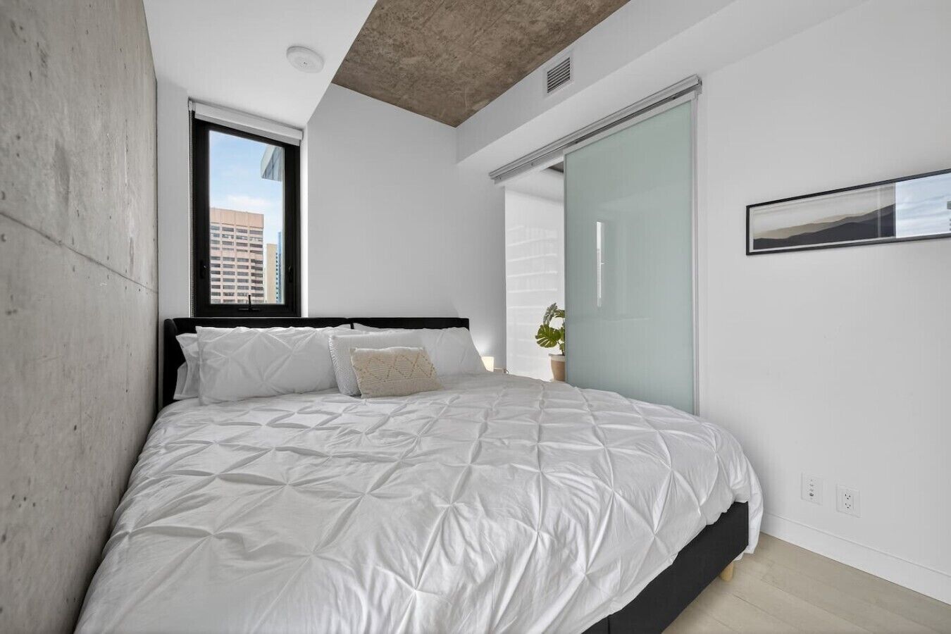 A modern bedroom with a large bed, a window, and a view of buildings outside.