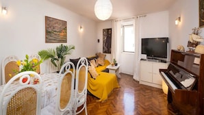 A living room with a wooden herringbone floor, a yellow sofa, a dining table with white chairs, a television, and a piano. - Apartment Jany (Dubrovnik)