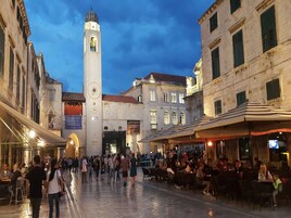 Uma praça movimentada com edifícios históricos, uma torre do relógio imponente e áreas de estar ao ar livre para cafés.