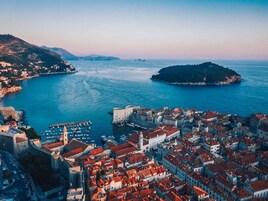 Vista aérea de uma cidade costeira com arquitetura histórica, um porto e uma ilha proeminente ao longe.