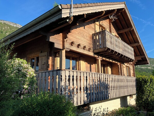 Un chalet en bois doté d’un balcon, entouré d’une végétation luxuriante et avec des montagnes en toile de fond.