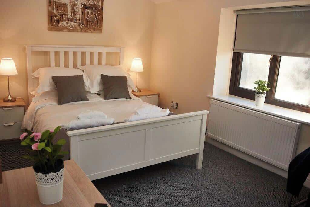A hotel room with a bed, two bedside lamps, a radiator, a window with a plant, and a painting on the wall.