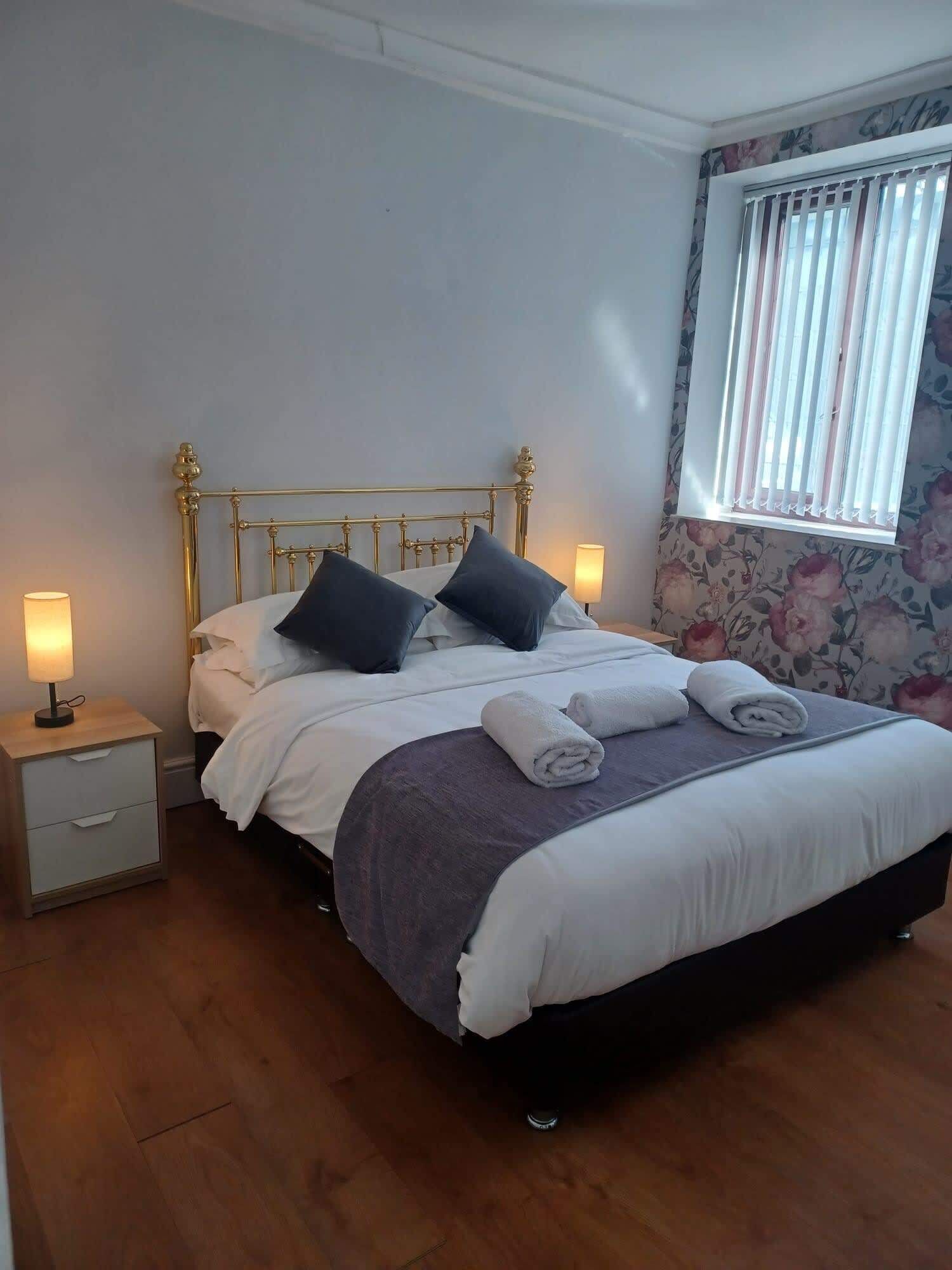 Een netjes opgemaakt bed met kussens, een nachtkastje met een lamp, en een raam met zonwering.