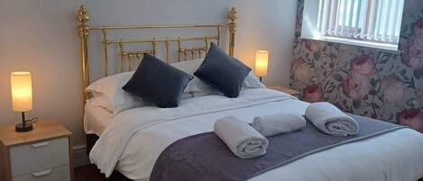 Een netjes opgemaakt bed met kussens, een nachtkastje met een lamp, en een raam met zonwering.