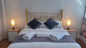 Een bed met een gouden hoofdeinde, twee nachtkastjes met lampen, en een grijs-witte spreidekker.