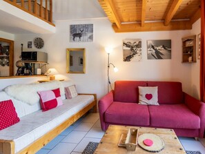 Ein gemütliches Wohnzimmer mit Holzbalkendecke, einem weißen Sofa mit roten und rosafarbenen Kissen, einem roten Sofa mit einem Herz-Kissen, einem Couchtisch mit Holzbeschlag und einem Spiegel an der Wand.