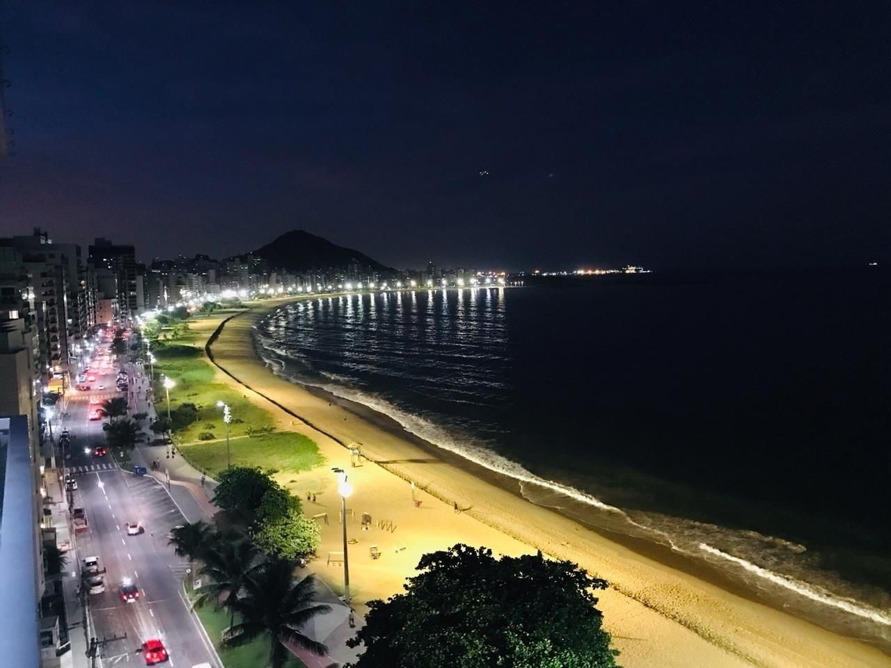 Ein Strandbild bei Nacht mit einer gut beleuchteten Promenade, einem Sandstrand und ruhiger See.