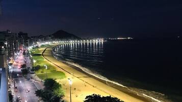 Ein Strandbild bei Nacht mit einer gut beleuchteten Promenade, einem Sandstrand und ruhiger See.