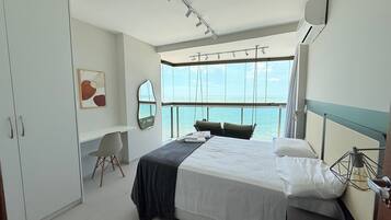 Quarto de hotel com cama, cadeira, escrivaninha, janela grande com vista para o mar e espelho na parede.