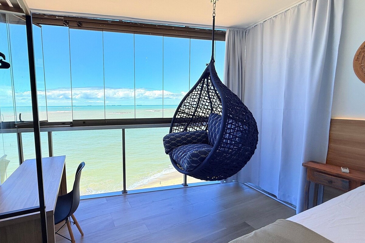 A room with a large window overlooking a beach, a hanging chair, a wooden table, and a bed.