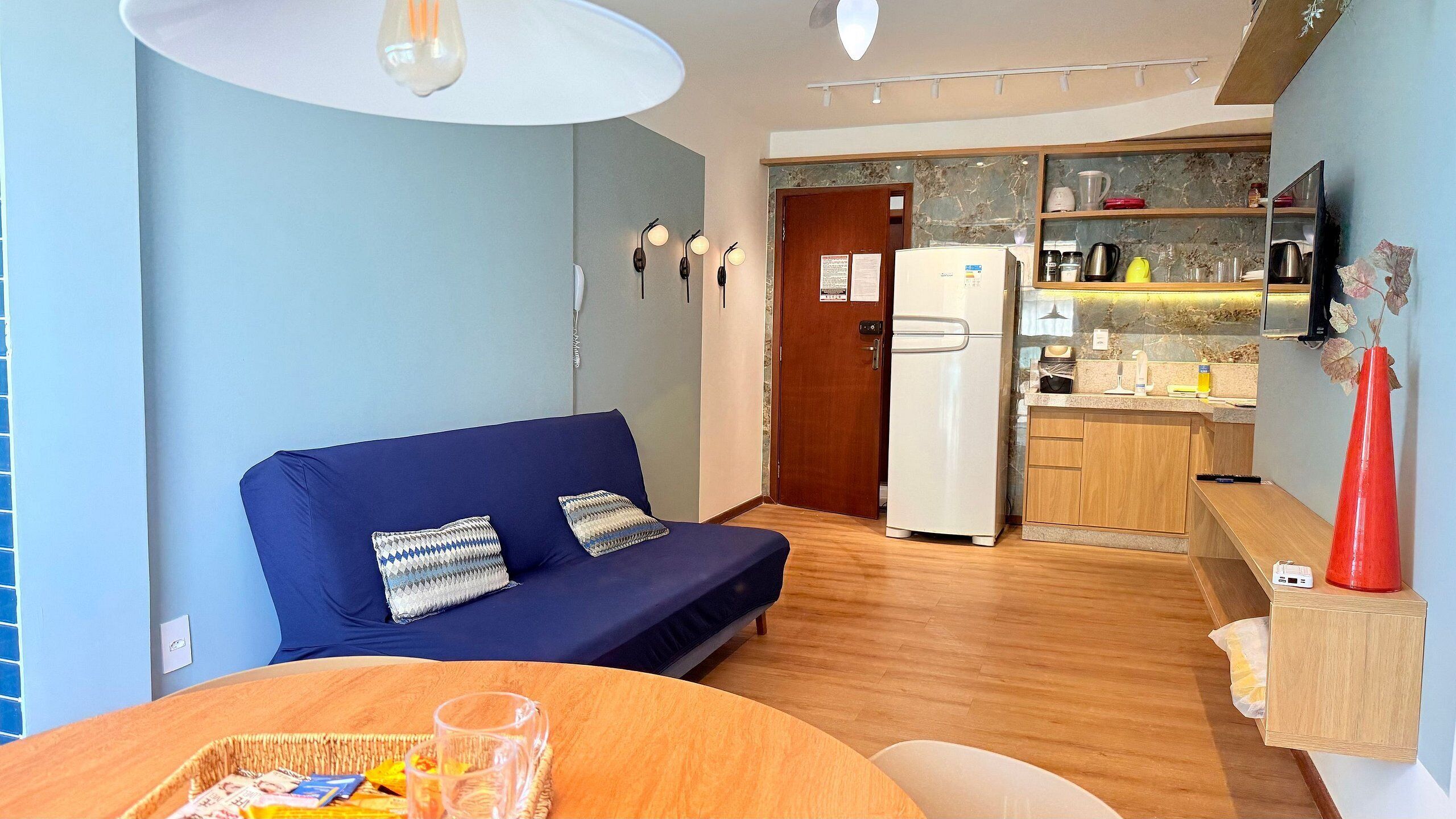 A compact living space with a sofa, a dining table with a basket, and a kitchen area with a refrigerator and various appliances.