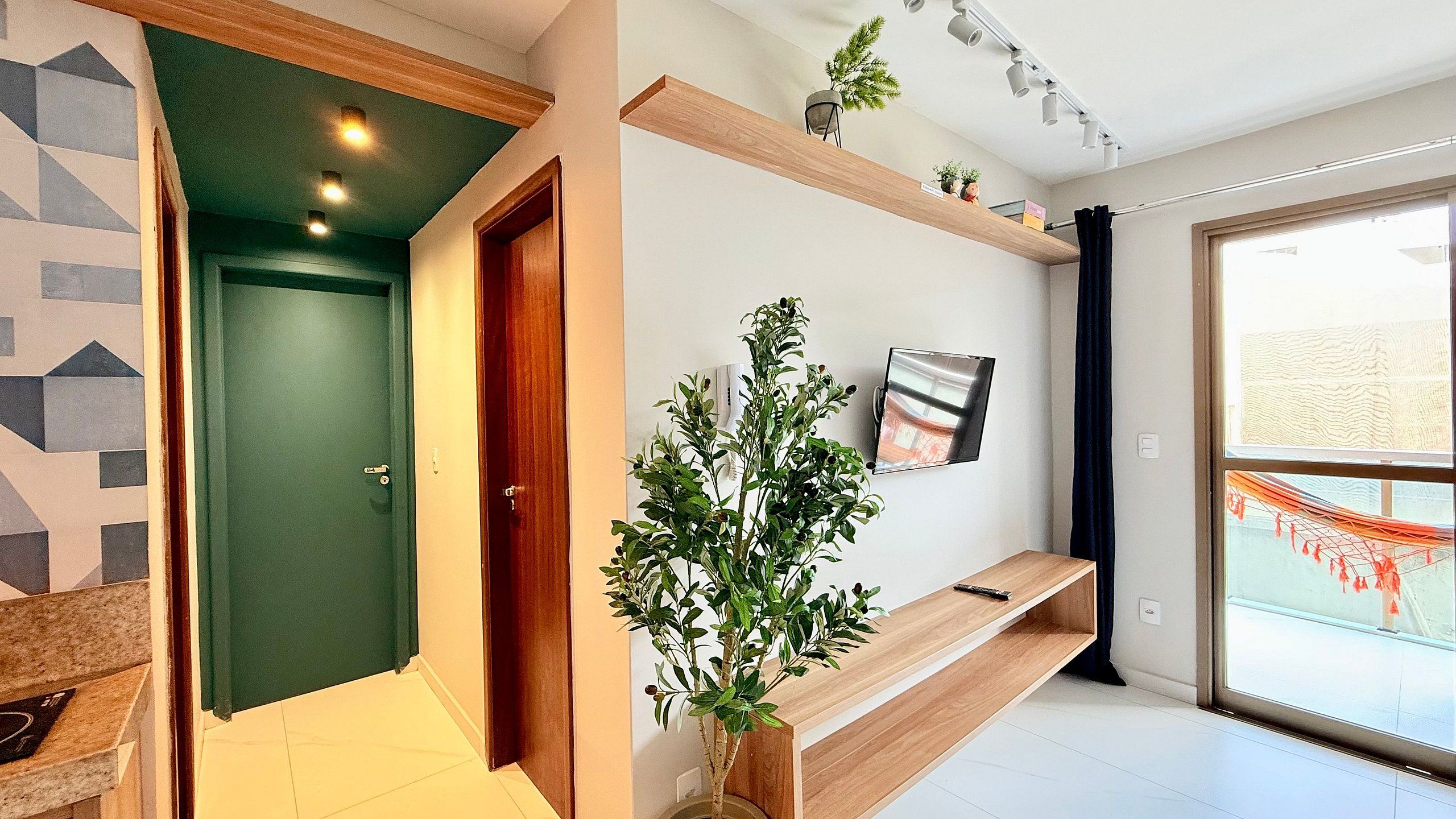 A modern hallway with a green door, a wooden bench, a potted plant, and a mounted TV.