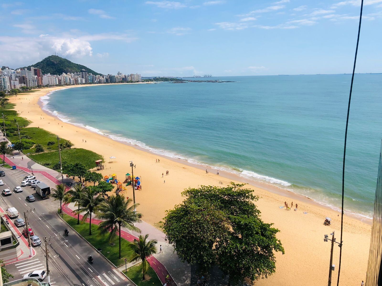 Ein Strand mit einer entlangführenden Straße, Palmen und einer Stadtansicht im Hintergrund.