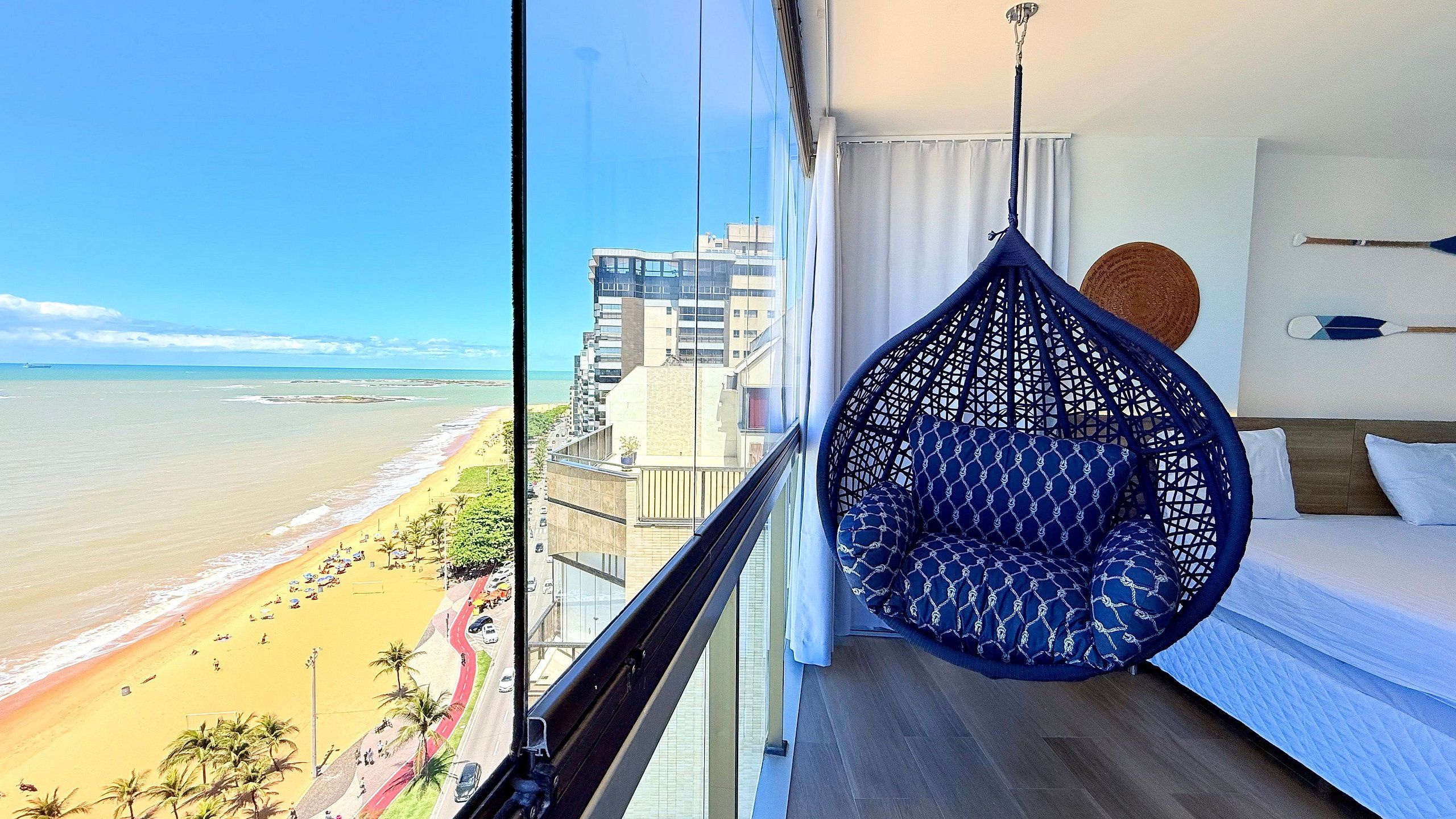 A balcony with a hanging chair, a bed, and a view of a beach and buildings.
