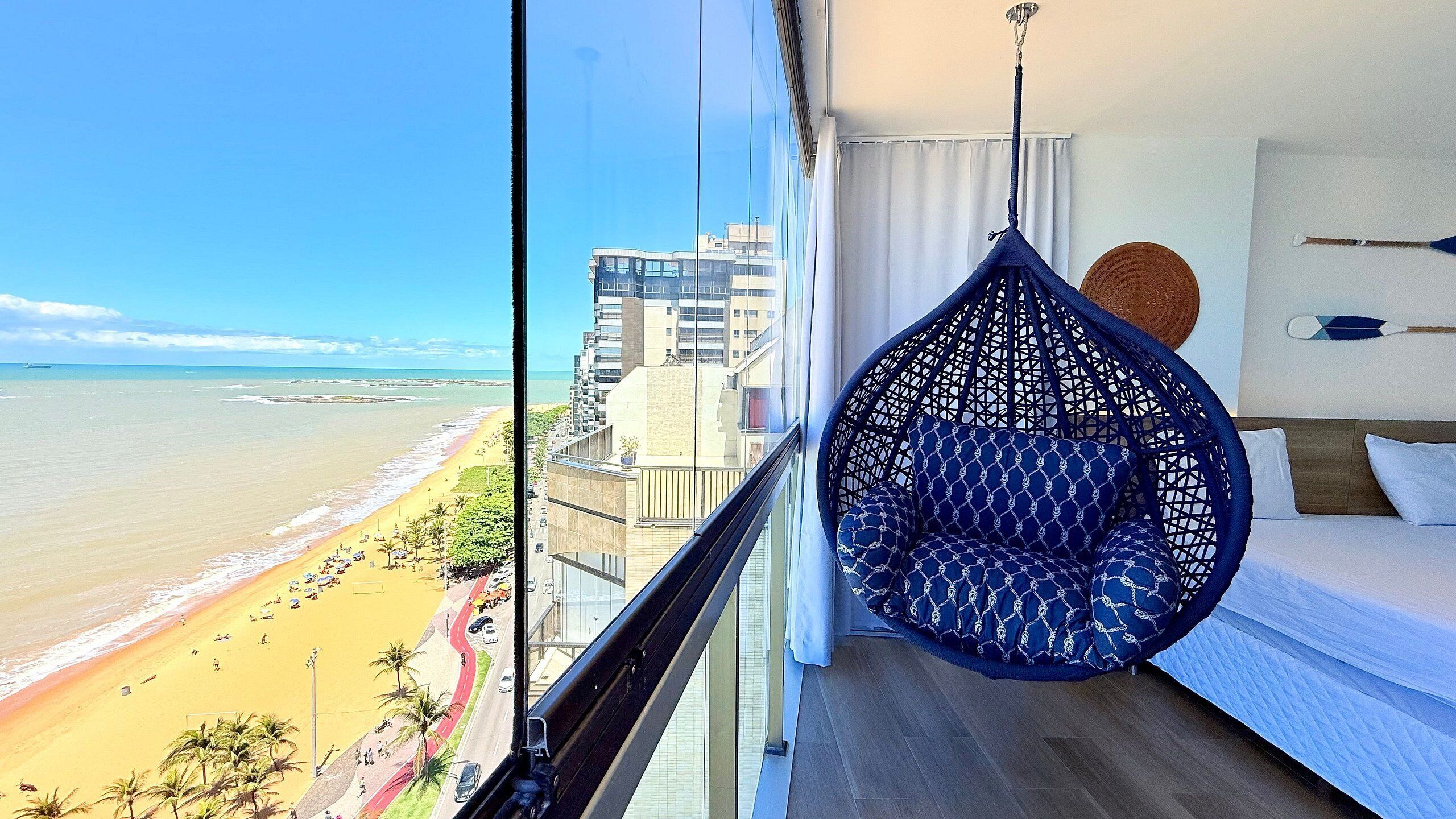 Uma varanda com uma cadeira suspensa, uma cama e vista para a praia e edifícios.