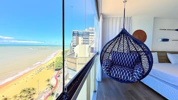 Uma varanda com uma cadeira suspensa, uma cama e vista para a praia e edifícios.
