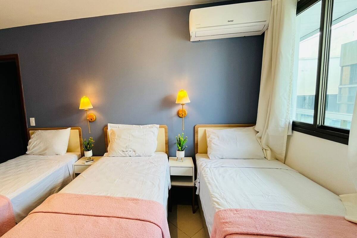 Habitación de hotel con tres camas individuales, una pared de color oscuro, un equipo de aire acondicionado y una ventana con cortinas.