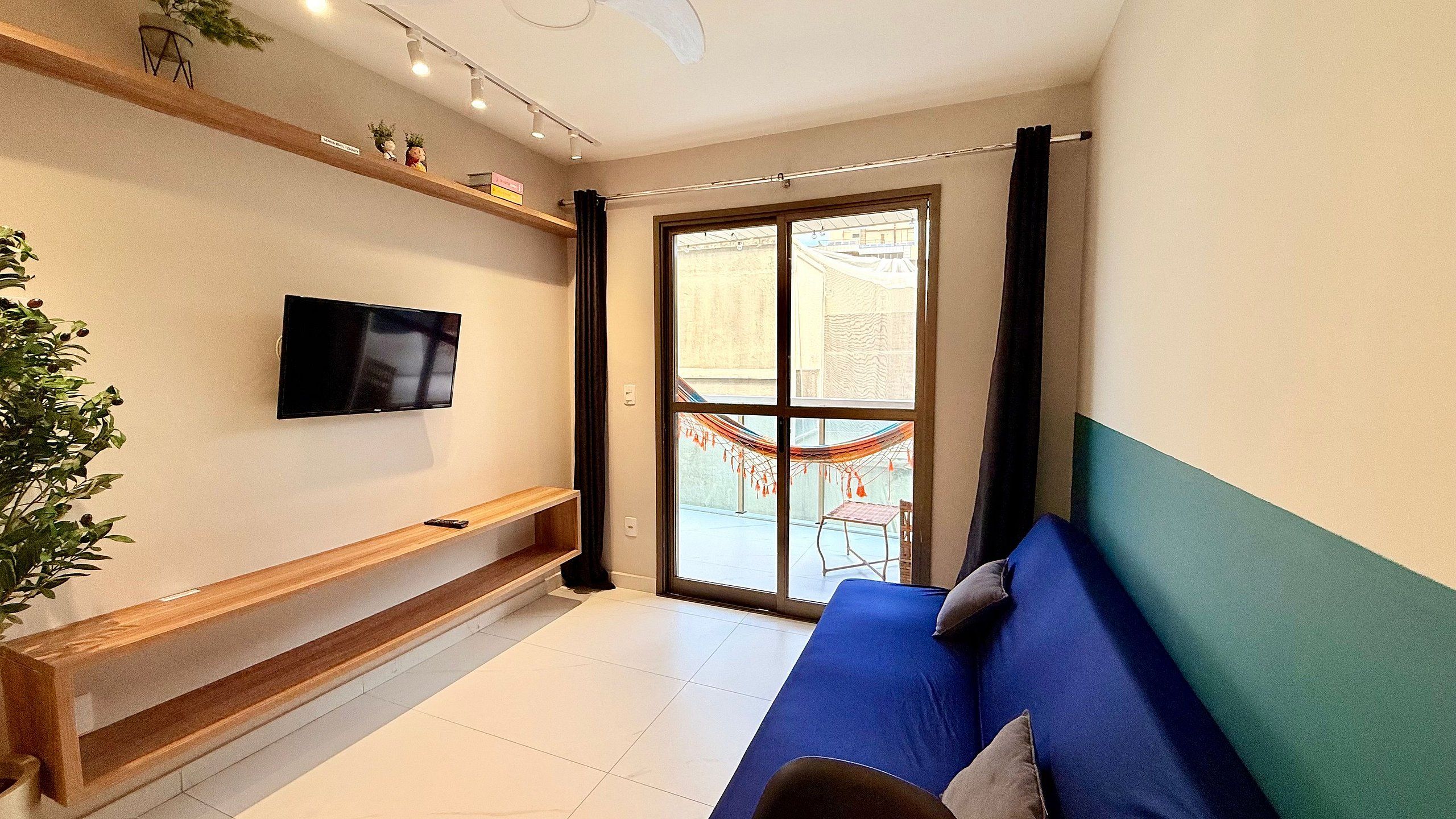 A modern living room with a flat-screen TV mounted on the wall, a blue sofa with cushions, a wooden bench, and a large sliding glass door leading to an outdoor area with a hammock.