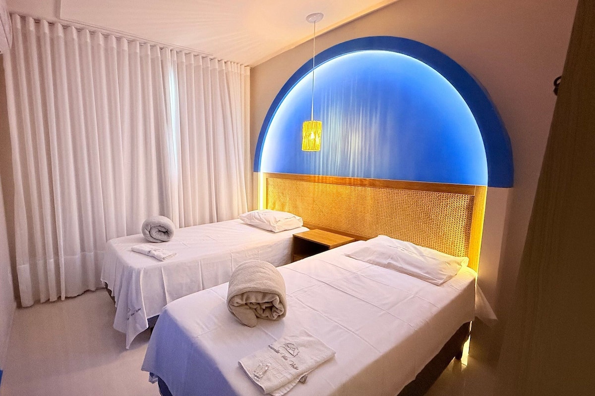 Habitación de hotel con dos camas, ropa de cama blanca, una lámpara azul en arco grande y cortinas blancas.