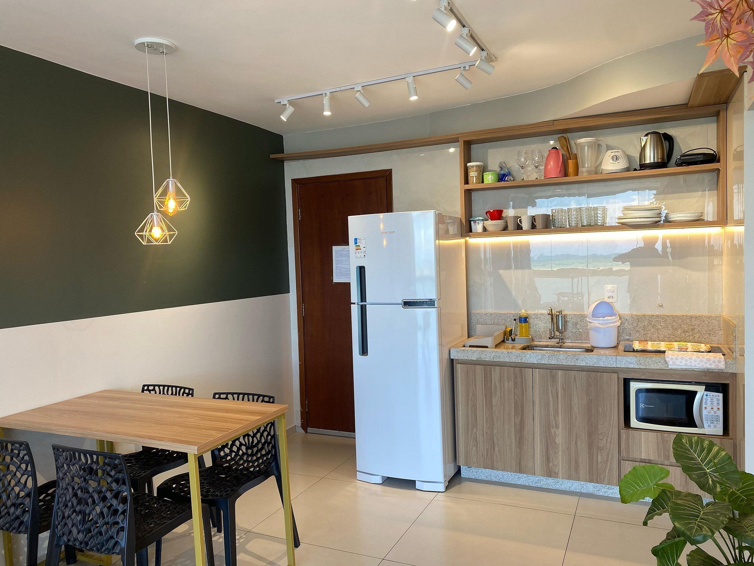A modern kitchen with a refrigerator, microwave, and wooden cabinets. There is a dining table with chairs and pendant lighting.